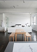 Pale wooden table and benches on grey concrete floor in front of white sleeping area in loft-style interior