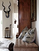 Scatter cushions on ottoman next to antique cupboard; hunting trophy on wall in background