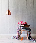 Stack of cushions on wooden chair and pendant lamp with copper lampshade in front of wooden wall
