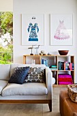 Comfortable designer sofa with scatter cushions, framed fashion illustrations and wooden shelves against wall in background