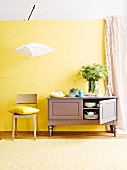 Crockery in small, country-house sideboard, modern wooden chair and Japanese rice paper lamp against yellow wall