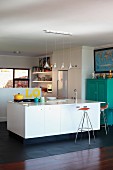 Row of pendant lamps above island counter with sink and bar stools; turquoise cupboard to one side in open-plan kitchen