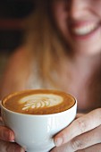 Von Frauenhänden gehaltene Tasse Cappuccino mit Milchschaummuster; unscharf im Hintergrund ein lachendes Frauengesicht