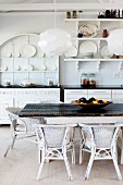 Modern pendant lamps above rustic table and white rattan chairs in front of kitchen counter and white crockery on wall-mounted shelves