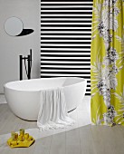 Towel hanging over edge of free-standing white bathtub amongst hanging lengths of fabric and small, round mirror on wall