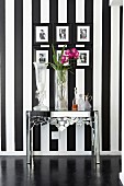 Various vases on chromed metal table below gallery of framed photos on black and white striped wall
