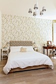Double bed with upholstered headboard against floral wallpaper and antique dressing table a small distance away