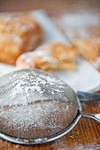 A sieve with icing sugar, baked pastries in the background