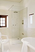 Partially visible white Ghost chair and bathtub in front of floor-level shower with glass partition in bathroom with white wall tiles