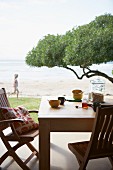 Zwei Holzstühle und Tisch mit Frühstücksutensilien auf Terrasse mit Blick auf Baumkrone und Meeresstrand