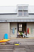 Holzterrasse mit Paddel und Boot vor Strandhaus mit Blechdach im Industriestil; spielende Kinder und Badeutensilien im offenen Flurbereich