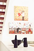 Still-life arrangement of black ceramic vessels on half-height cabinet in front of niche below staircase; pictures with motifs of children on wall