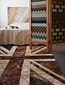 Geometrische Patchworkmuster im Schlafzimmer für Lederteppich und Wandbehang hinter dem Doppelbett; in der Mitte ein Paravent mit stilisiertem Webmuster