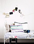 Scatter cushions on metal bed with curved, white frame below collection of postcards hanging from looped cord on wall