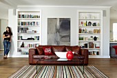 Plexiglass coffee table on striped rug and leather couch in front of fitted shelving; woman in background