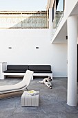 Shady terrace of modern town house with grey stone tiles, bench with black seat cushions and white designer lounger