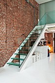 Staircase with glass balustrade and glass steps against exposed brick wall in designer loft apartment with white floor; map cabinets below stairs