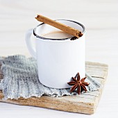 weiße Emaille Tasse mit Tee, darauf liegt eine Zimtstange und ein Anisstern lehnt an der Tasse, alles steht auf einem gestrickten grauen Flicken und einem verwittertem Holzbrett