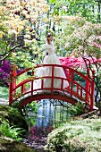 Braut in weißem Brautkleid auf roter Brücke in Parkanlage