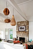 Fishing hut converted into weekend cottage with exposed, white-painted roof and pendant lamps with wooden lampshades above seating area with stone chimney breast
