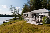 Zu Wochenendhaus ausgebaute Fischerhütte im weissen Long- Island-Stil am See; Plattform mit Liegestühlen und überdachte Eingangsveranda