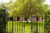 Open wrought iron gate leading to garden with tree and English house with brick facade