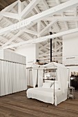 Four-poster bed with white fabric canopy in sleeping area below white roof beams of loft apartment
