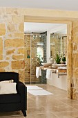 Durchgang in Natursteinmauer mit Blick auf Waschtisch und Dusche in modernem Bad; Sessel im Vordergrund