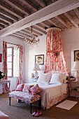 Romantic, shabby-chic bedroom with red and white Toile de jouy fabric curtains, bedroom bench, scatter cushions and canopy below rustic, wood-beamed ceiling