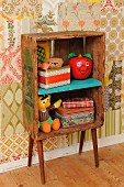 Retro-style child's bedroom with DIY shelves made from old wooden crate and 50s cabinet legs; old tin, vintage Bert and Happy Apple toys against patchwork of 70s wallpaper
