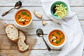 Two Bowls of Gazpacho Garnished with Cilantro Oil, Chopped Cucumbers and Dried Chili Peppers; Served with Bread