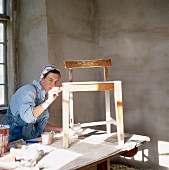 Frau mit Latzhose beim Lackieren eines Holzstuhls auf mit Zeitungspapier abgedecktem Tisch