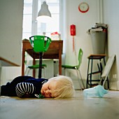 Toddler asleep on kitchen floor with dropped-out dummy; bright green suspended highchair at dining table in background