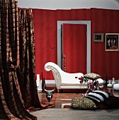 Sophisticated living room with white chaise longue and elegant cushions on floor in front of red wallpapered wall with lengths of fabric draped to one side