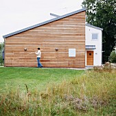 Rasenfläche vor modernem Wohnhaus und Mann mit Gartengerät vor Holzfassade