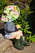 Toddler wearing sun hat and wellingtons sitting on stone step in garden