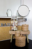 Small arrangement of woven containers and collection of shells against mosaic tiles; black washstand, mirrored cabinet and magnifying mirror on jointed arm