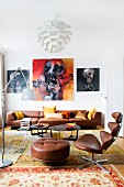 Classic office chair with brown leather cover and footstool in front of lounge area with black coffee table and brown leather sofa below expressive paintings on wall