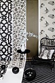 Black disco balls, side table, wicker chair and scatter cushion with print portrait in front of lengths of patterned wallpaper