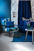 Seating area in shades of blue with armchair, sofa and side able; display case of mounted butterflies on patterned wallpaper