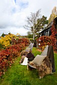 Zwei Gartenstühle mit wärmenden Felldecken und Kissen in einem mit herbstlichem Weinlaub berankten Garten