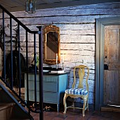 Schmiedeeisernes Treppengeländer; dahinter eine blaue Kommode mit goldgerahmtem Spiegel an rustikaler Holzwand