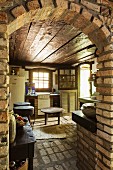 Blick durch gemauerten Rundbogen in rustikale Küche mit dunkler Holzdecke, Ziegelboden und Klinkerwänden