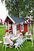 Kinder sitzen an Tisch vor Spielhaus im Garten