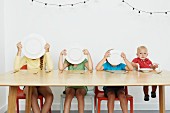 Four children sitting at table, three covering faces with plates