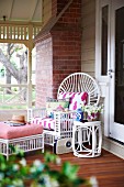 White, wicker peacock chair with seat cushion, scatter cushions, matching side table and footstool with cushions on veranda