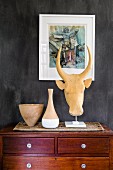 Clay pots and animal head sculpture on chest of drawers below small painting on dark wall