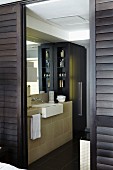 Junggesellen Bad Ensuite hinter geöffneten dunkelbraunen Schiebejalousietüren, eleganter Waschtisch mit schwarzem Einbauschrank
