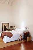 Corner of minimalist bedroom - dog sitting on sisal rug in front of double bed with white bed linen, retro table lamp on bedside table, picture on floor leaning against wall