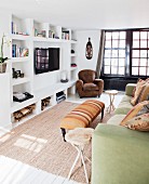 Sofa with scatter cushions and ottoman opposite wall with masonry shelving and wall-mounted TV; leather armchair in background next to black, latticed French doors